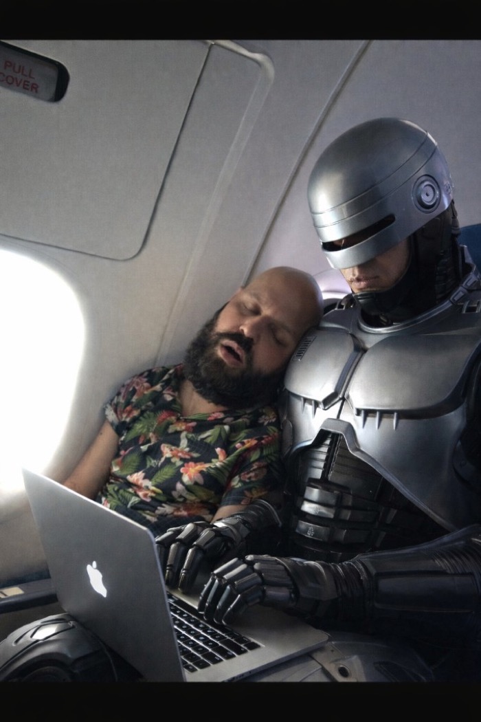 Robocop coding on a MacBook while a man sleeps on his shoulder in an airplane cabin